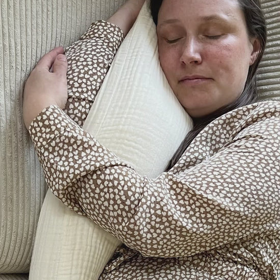 Kvinde slapper af med Kadera Mommy Pillow i cream på sofaen, hvor puden giver blød og behagelig støtte.